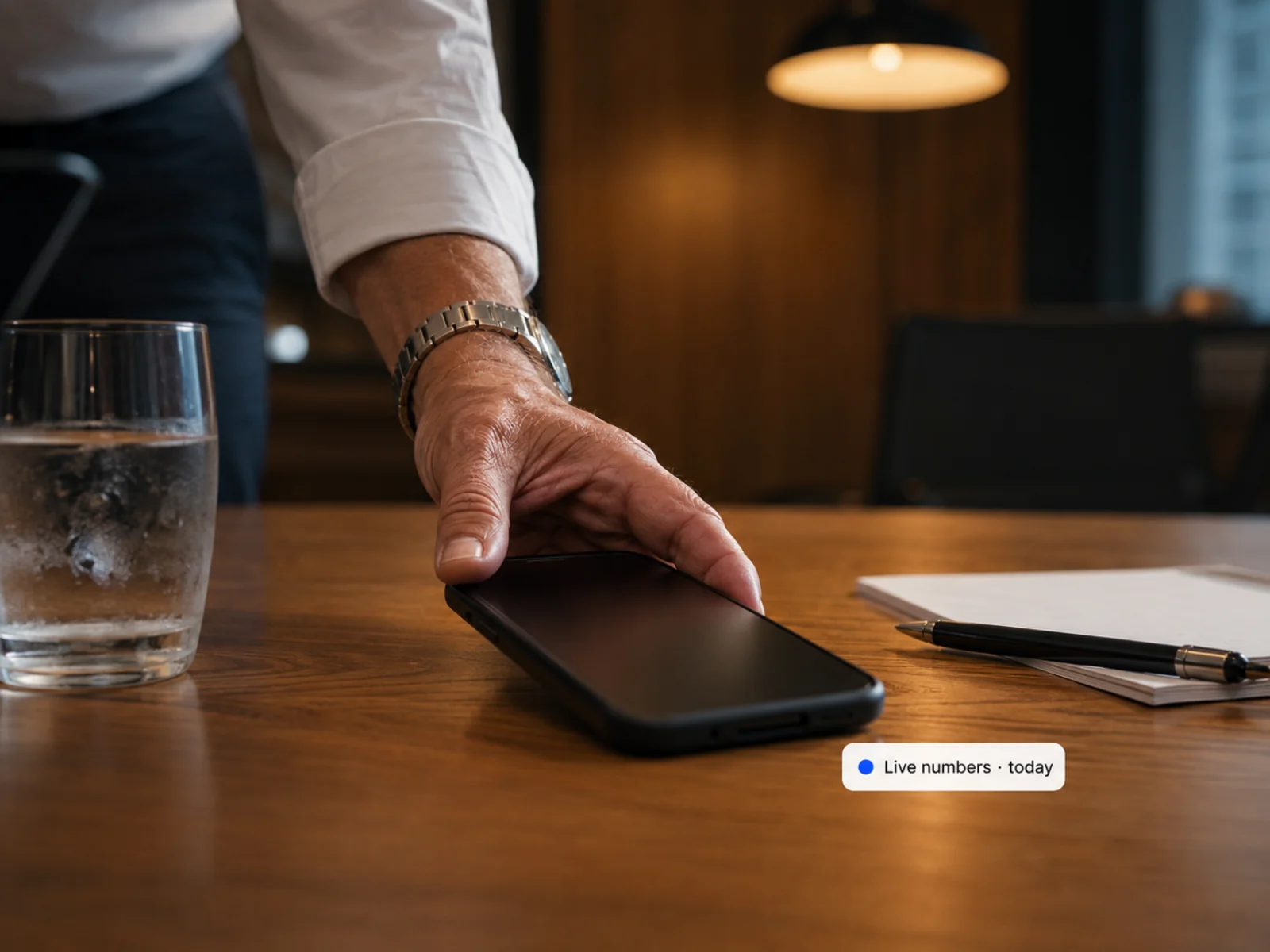A hand sliding a phone across a meeting table with a live-numbers-today chip